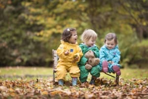 Three small children wearing puddle suits and wellies, sitting on a bench in a park surrounded by brown autumn leaves and trees in the background.