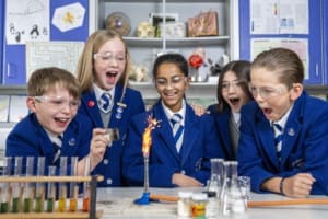 Five junior school pupils from Kelvinside Academy wearing blue blazers and science goggles, leaning on a workshop table, looking surprised and excited by a Bunsen burner alight. They are surrounded by science materials like test tubes, flasks, & diagrams.